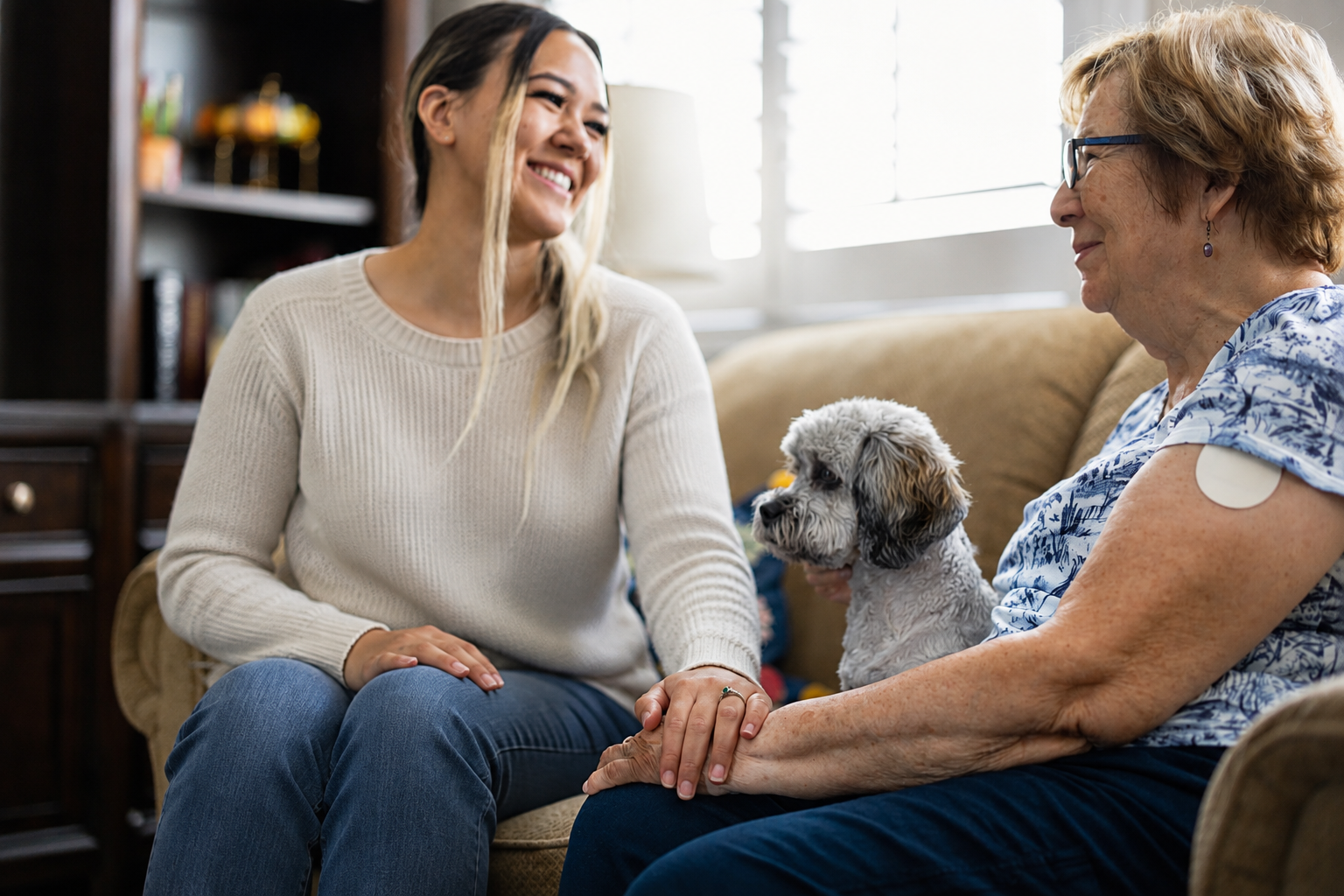 Young woman in casual clothing providing companionship and support to an older woman at home, with a small dog sitting between them.